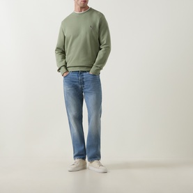 Model wearing sage green crew neck sweatshirt with small chest logo, styled with blue straight-leg jeans and light grey trainers.