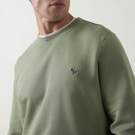 Model wearing sage green crew neck sweatshirt with embroidered zebra logo badge on chest, layered over white crew neck tee