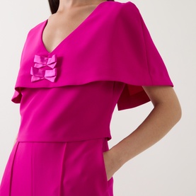 Model wearing Sisters fuchsia jumpsuit with cape overlay and satin double bow detail at chest.
