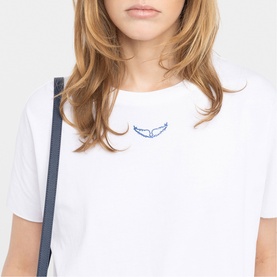 Model wearing Zadig white T-shirt with blue embroidered wing motif on chest, styled with a dark shoulder bag strap visible.