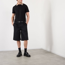 Model wearing Burberry black cotton polo shirt with check collar detail, styled with black shorts, belt, socks and chunky loafers