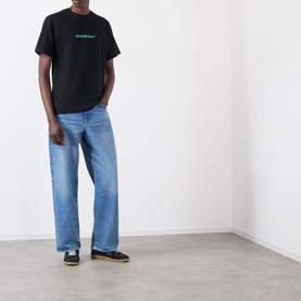 Model wearing Burberry Ryan Big Logo T-Shirt in black with green logo print, styled with light blue wide-leg jeans and dark trainers.