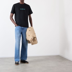 Model wearing Burberry Ryan Big Logo T-Shirt in black with blue jeans and dark trainers, carrying a tan backpack.