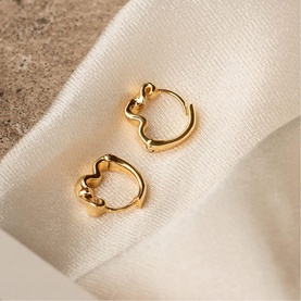 Daisy Jewellery Est&eacute;e Lalonde Flow Huggies in 18ct gold plate, close-fitting huggie hoops with smooth twisted wave detail, styled on ivory fabric