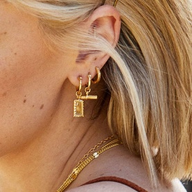 Model with Daisy Jewellery Est&eacute;e Lalonde T-Bar Charm Earrings in gold tone, styled with stacked hoop earrings and layered chain necklaces.