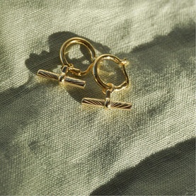 Daisy Jewellery Est&eacute;e Lalonde T-Bar Charm Earrings in gold-tone, featuring small drop hoops with engraved T-bar charms, on sage fabric