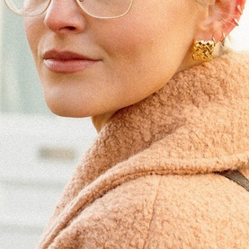 Model with Daisy Jewellery Polly Mini Woven Square Studs in 18ct gold plate, textured square silhouette. Styled with pink teddy coat and glasses.