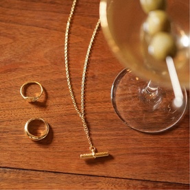 Daisy Jewellery Est&eacute;e Lalonde T-Bar Necklace in 18ct gold plate, with engraved T-bar pendant on fine chain, styled on wood surface with gold rings