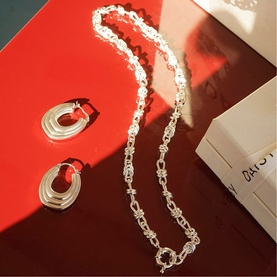 Daisy Jewellery Polly Knot Chain Necklace in sterling silver with bold knot links, styled alongside silver chunky hoop earrings on red surface.