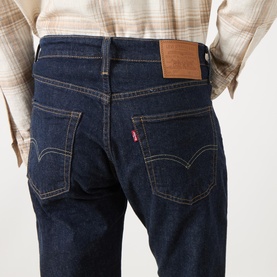 Model wearing Levi's dark blue jeans, rear view showing iconic red tab, leather patch, and contrast stitching. Styled with a checked shirt.
