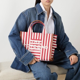 Model carrying Marc Jacobs The Striped Canvas Medium Tote Bag in True Red Multi, red leather top handles, bold logo print, styled with denim jacket