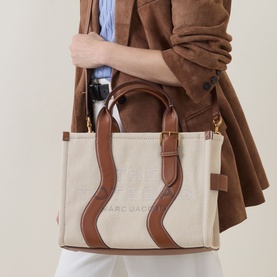 Model carrying Marc Jacobs The Wavy Canvas Medium Tote Bag in beige/argan oil with brown leather handles, wavy contrast trim and gold-tone hardware.