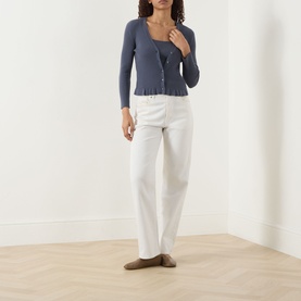 Model wearing Paige Pacific Blue cardigan with scoop neck and frill hem, styled with white wide-leg trousers and taupe flats.