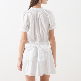 Model wearing Paige white cotton mini dress, rear view, featuring puff sleeves, gathered neckline, and tie-back waist detail with tiered skirt
