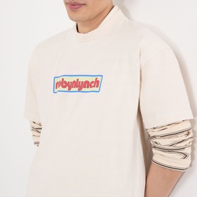 Model wearing Robyn Lynch Logo Graphic T-Shirt in off white with blue and red chest logo, styled over a long-sleeve top.