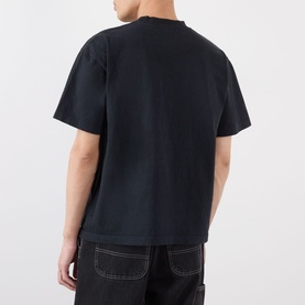 Model wearing Robyn Lynch Skint Logo T-Shirt in off black, rear view, dropped shoulders, straight hem, styled with black contrast-stitch trousers.