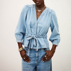 Model wearing Bash blue denim peplum blouse with V-neck, puff sleeves, and tie waist, styled with wide-leg jeans and gold jewellery