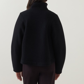 Model wearing Annette Gortz black cotton jacket, rear view, with turtleneck collar and relaxed boxy silhouette, styled with burgundy trousers.