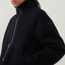 Model wearing Annette Gortz black zip-up jacket with high funnel neck, silver zip detail, close-up crop shot.