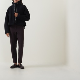 Model wearing Annette Gortz black cotton high-neck jacket with dark tapered trousers and brown loafers, long pendant necklace