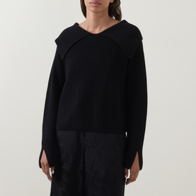 Model wearing Annette Gortz black merino wool knit jumper with draped collar detail, styled with black wide-leg trousers