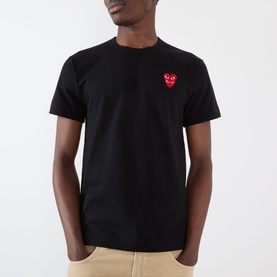 Model wearing Comme des Gar&ccedil;ons Play black cotton T-shirt with signature red double heart logo patch on chest, styled with beige trousers.