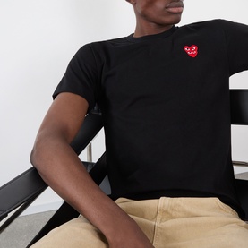 Model wearing Comme des Gar&ccedil;ons Play black cotton T-shirt with red double heart logo badge, styled with tan trousers.