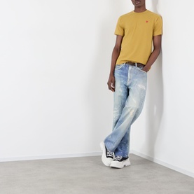 Model wearing Comme Des Gar&ccedil;ons Play olive cotton T-shirt with red heart logo, styled with light wash distressed jeans and black trainers.