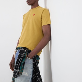 Model wearing Comme des Gar&ccedil;ons Play olive cotton T-shirt with small red heart logo, styled with distressed jeans and plaid shirt tied at waist.