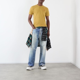 Model wearing Comme Des Gar&ccedil;ons Play olive cotton T-shirt with red heart logo, styled with light wash jeans and plaid shirt tied at waist.