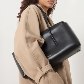 Model carrying DeMellier Stockholm Shoulder Bag in black grained leather with gold-tone hardware and tubular handles, styled with a camel oversized