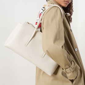 Model carrying DeMellier Stockholm Shoulder Bag in chalk grained leather, gold-tone hardware, top zip fastening, styled with a beige trench coat and