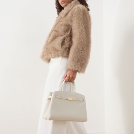 Model carrying DeMellier The Hudson Midi Top Handle Bag in Chalk grained leather with gold-plated hardware, styled with a beige faux fur jacket and