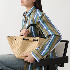 Model carrying DeMellier The New York Midi Top Handle Bag in warm raffia and black with tubular handles, styled with a striped shirt.