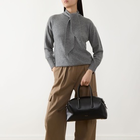 Model carrying DeMellier The Stockholm Midi Top Handle Bag in black grained leather with gold hardware, styled with grey knit and tan trousers.
