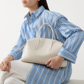 Model carrying DeMellier The Stockholm Midi Top Handle Bag in Chalk grained leather with gold-tone hardware and tubular top handles, styled with a