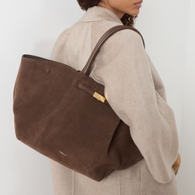 Model carrying DeMellier The New York Suede Top Handle Bag in mocha suede with gold-plated hardware, worn over shoulder, styled with a beige coat.
