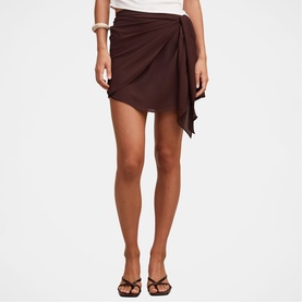 Model wearing dark brown draped wrap mini skirt with asymmetric hem and tie waist, styled with brown strappy sandals