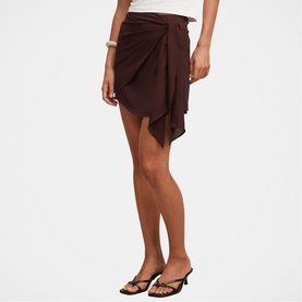 Model wearing dark brown draped wrap mini skirt with asymmetric hem, styled with white top and strappy heeled sandals