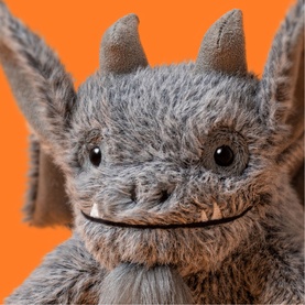Jellycat grey bat soft toy close-up, showing fluffy textured fur, small horns, beady eyes and a wide smile, against orange background