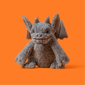 Jellycat grey fluffy dragon plush toy with wings, horns, and a grinning expression, shown against an orange background