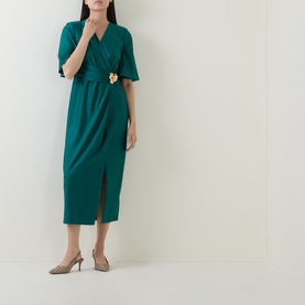 Model wearing Joseph Ribkoff Dress in Opulent Green, wrap-style midi with flutter sleeves and gold floral brooch, paired with taupe heels