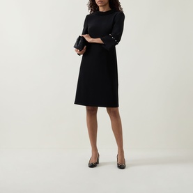 Model wearing Joseph Ribkoff Jewel Embellished Dress in black, midi shift silhouette with jewel-embellished cuffs, styled with black clutch and pumps.
