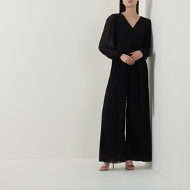 Model wearing Joseph Ribkoff black pleated wide-leg jumpsuit with V-neckline and long blouson sleeves, styled with strappy heeled sandals.