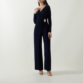 Model wearing Joseph Ribkoff Jumpsuit in Midnight Blue, long sleeves, wrap-style bodice with gold brooch detail, wide-leg trousers, styled with nude