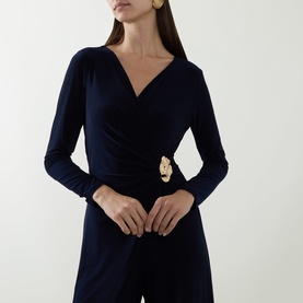 Model wearing Joseph Ribkoff Midnight Blue wrap-front jumpsuit with long sleeves and gold floral brooch detail at waist.