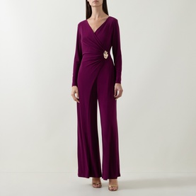 Model wearing Joseph Ribkoff Berry Burst jumpsuit with wrap-style V-neckline, long sleeves, gold brooch detail, and wide-leg trousers.