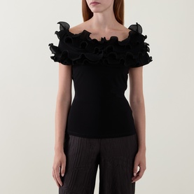 Model wearing Joseph Ribkoff black off-shoulder top with layered ruffle neckline, styled with black wide-leg trousers.