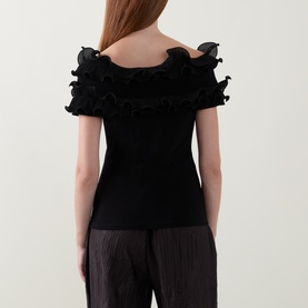 Model wearing Joseph Ribkoff black top with layered ruffle off-shoulder neckline, styled with grey pleated trousers. Rear view.
