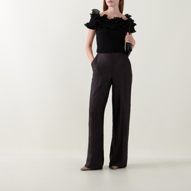 Model wearing Joseph Ribkoff black off-shoulder top with ruffle trim, styled with wide-leg black trousers and metallic pointed-toe flats.